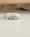 A set of stackable silver rings, with one clear cz ring and two personalized name rings, displayed on a wooden surface.