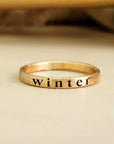yellow gold ring sitting on a light wood table. Engraved deeply on the ring in serif font, the name "winter". Shadows in background.