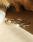 Two rings, one with a plain band and the other featuring a birthstone, displayed on a textured surface with a soft shadow.