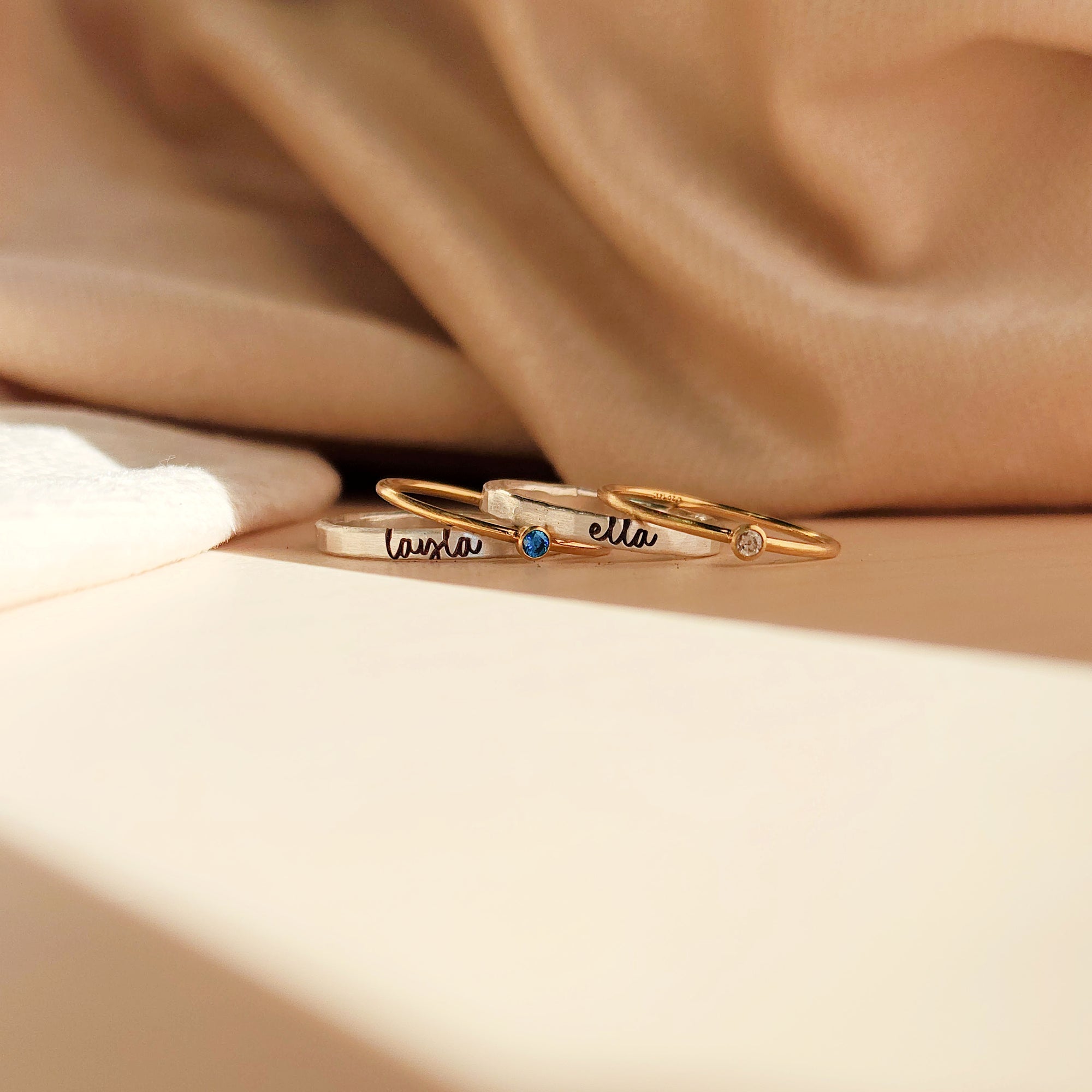 The rings include two thin gold bands with small gemstones and a slightly thicker silver band with engraved names in cursive. One gold ring has a blue gemstone and the engraved name 'laura,' while the other has a clear gemstone. The silver band in the center has the name 'ella' engraved.