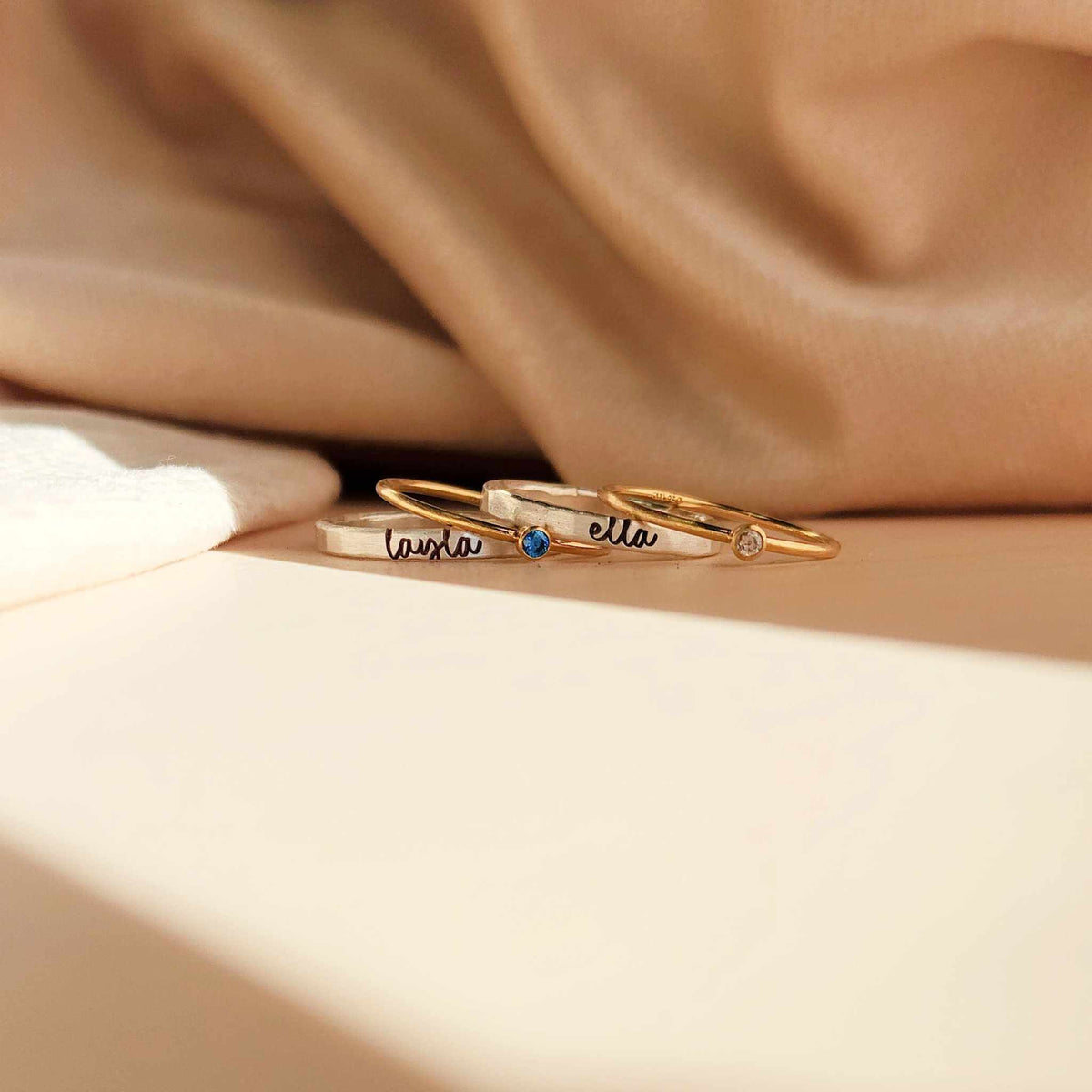 The rings include two thin gold bands with small gemstones and a slightly thicker silver band with engraved names in cursive. One gold ring has a blue gemstone and the engraved name 'laura,' while the other has a clear gemstone. The silver band in the center has the name 'ella' engraved.