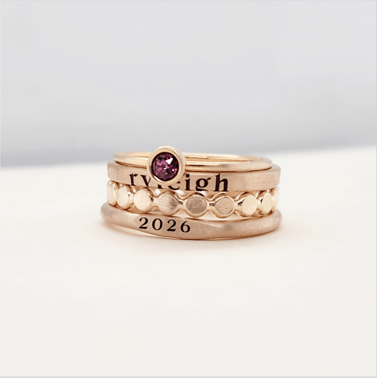 A set of stackable gold rings on a person's hand, featuring personalized engravings such as a name and the year 2026.