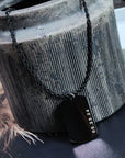 Black dog tag necklace with a date on it, hanging from a textured gray vase.