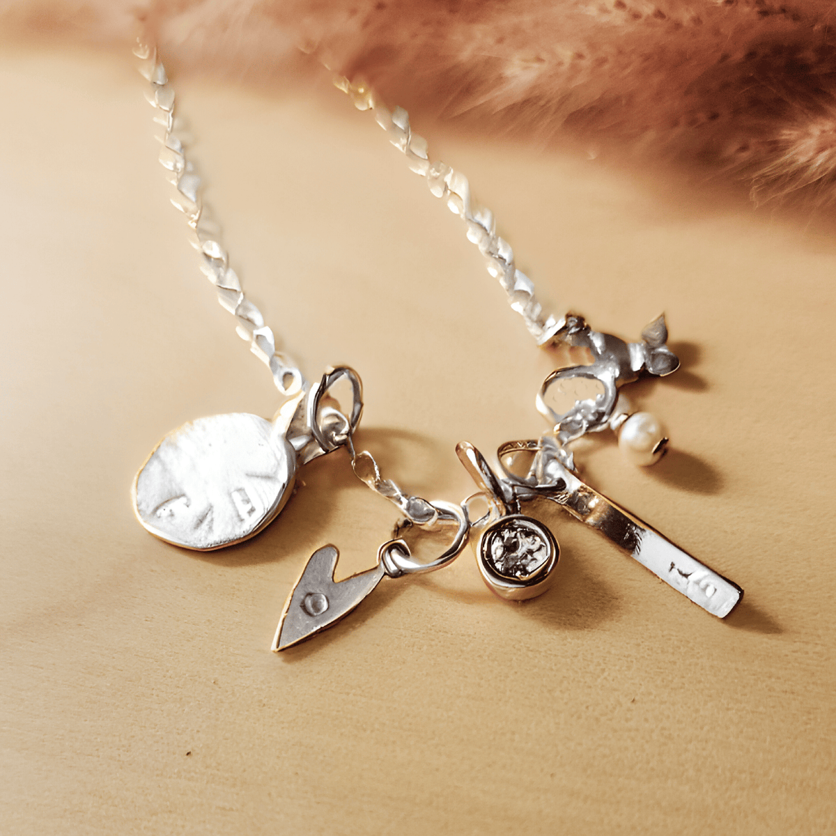 Close-up of a silver charm necklace displayed on a light wood surface with soft pink pampas grass in the background. The necklace features multiple silver charms, including: a round disc stamped with "GAB", a small heart with an engraved letter, a bezel-set clear crystal charm, a vertical bar engraved with "10", a small pearl dangle, and a delicate leaf charm. The chain is a shiny flat curb style, giving the piece a polished, customized look.
