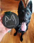 A black slate dog ear ornament with a velvet fringe ribbon, held in a hand, with a dog in the background.