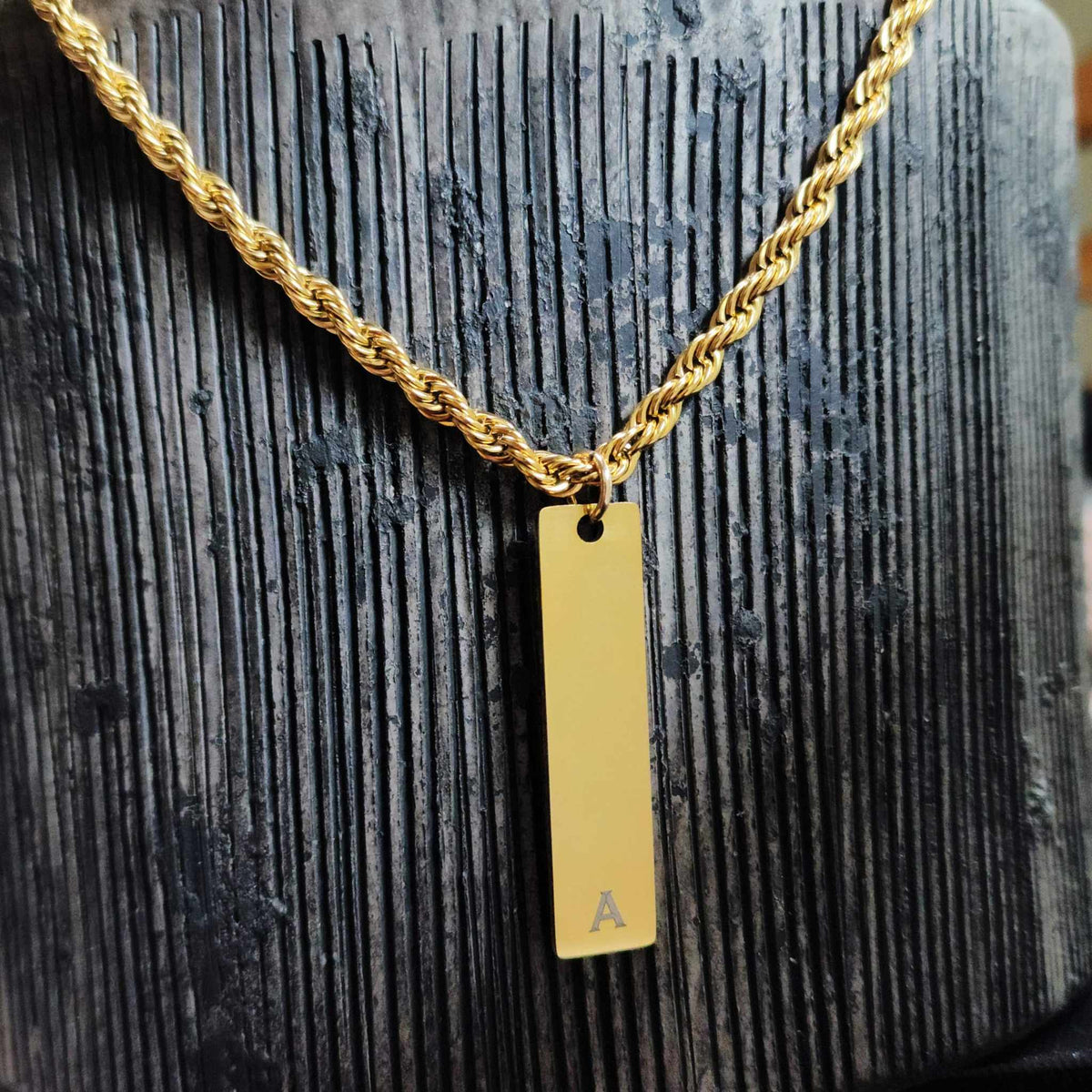 Gold necklace with a vertical bar pendant featuring the letter 'A' on a textured dark surface.