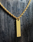 Gold necklace with a vertical bar pendant featuring the letter 'A' on a textured dark surface.