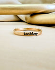 A gold-colored personalized stacking name ring with cursive writing on a textured band, placed on a beige surface with a reflective object in the background.