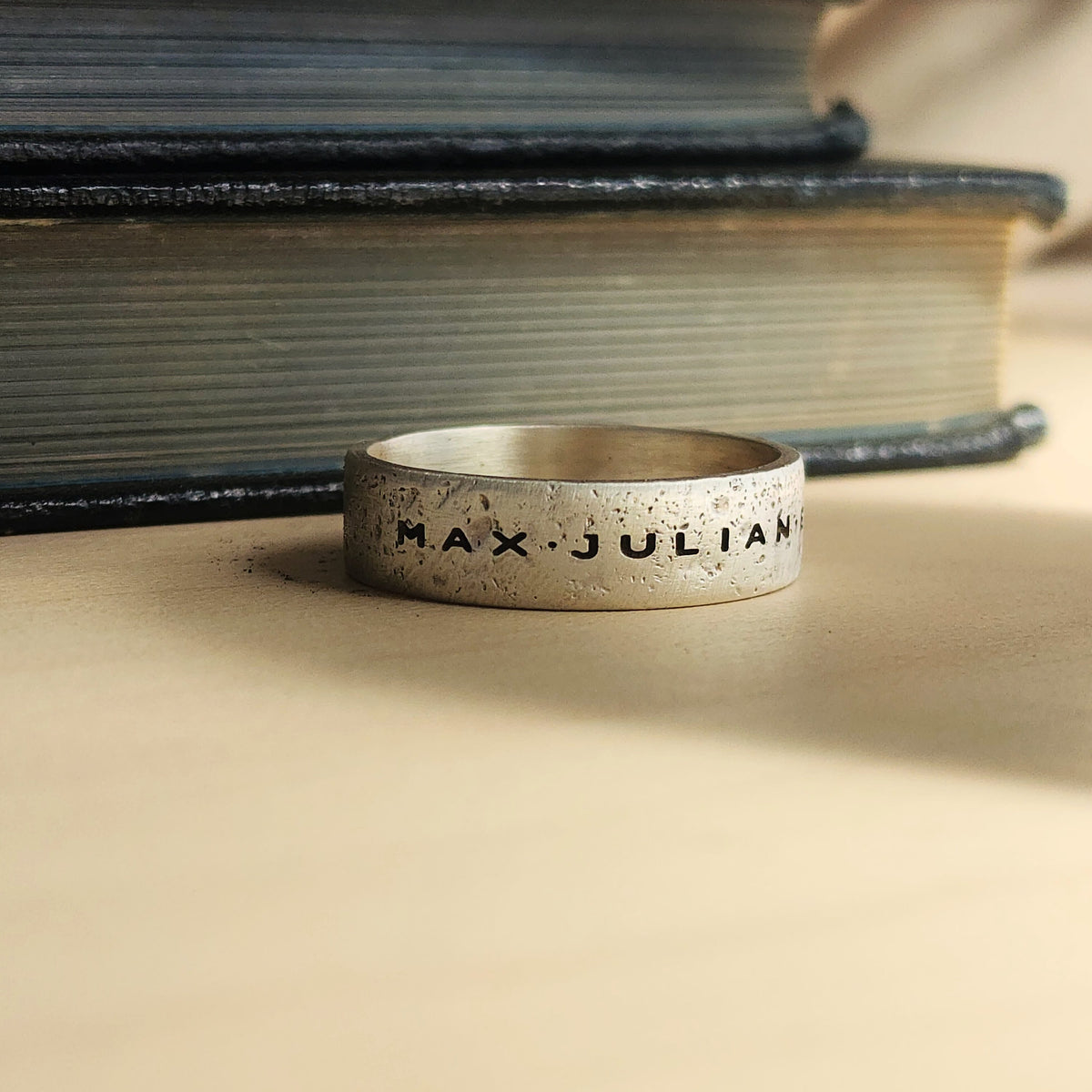 Silver ring with 'MAX JULIAN' engraving on a textured surface with books in the background