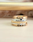 A set of mixed metal stacking name rings with personalized engravings on each ring, displayed on a person's hand.