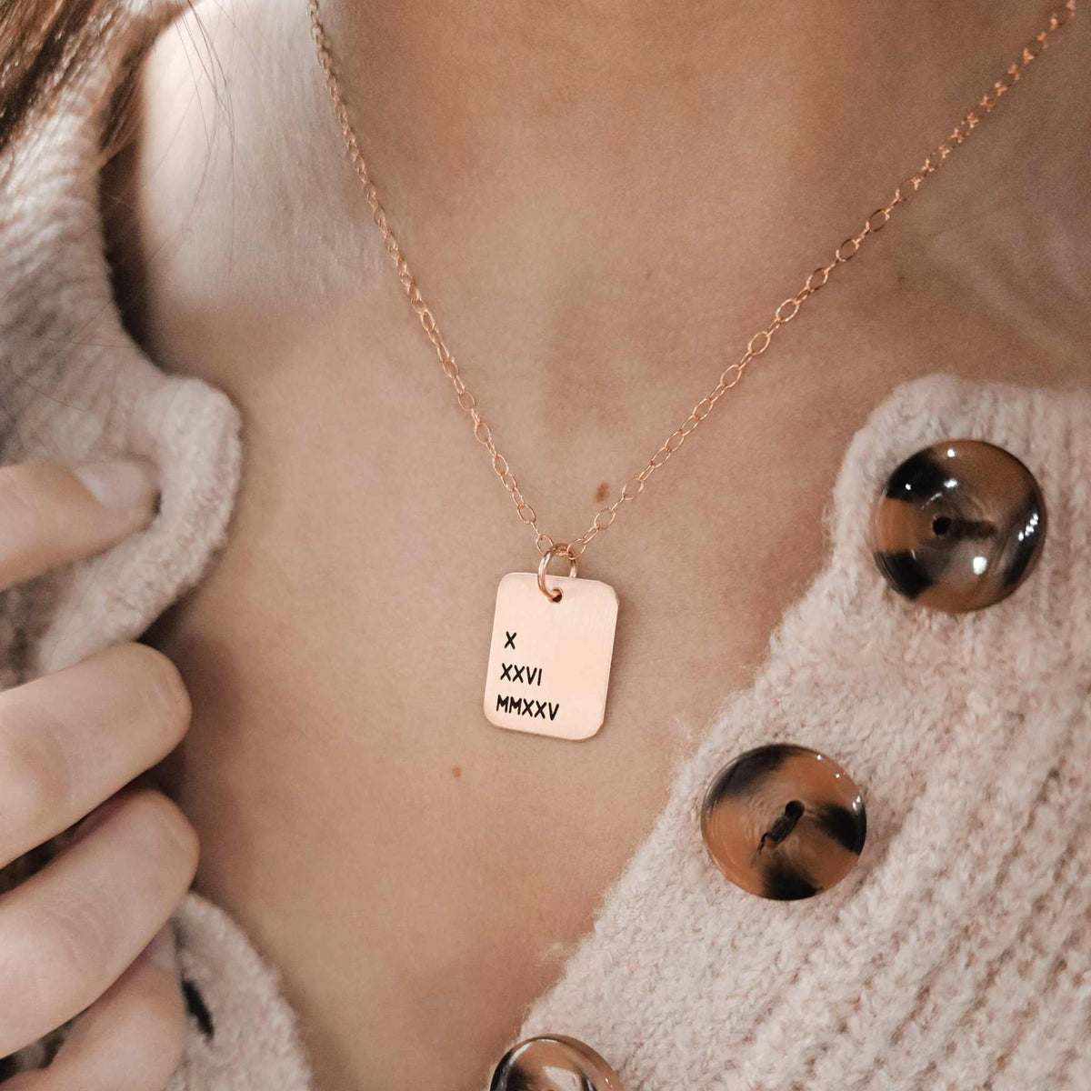 A person wearing a cardigan is showcasing a gold necklace with a rectangle pendant engraved with Roman numerals.