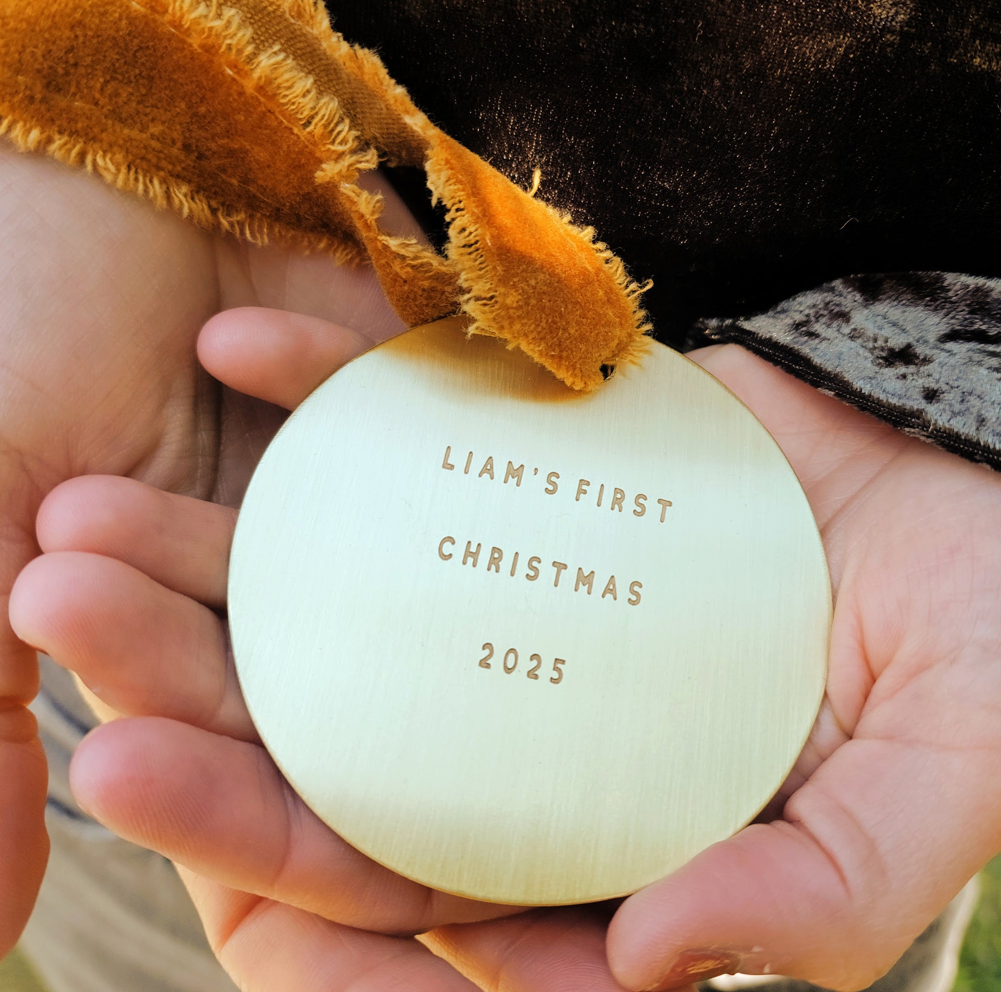 Child holding a 'Liam's First Christmas 2025' ornament with a teddy bear in the background.