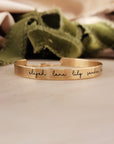 A gold-colored cuff bracelet with custom names engraved on it, displayed on a white surface with a green textured object to the side.
