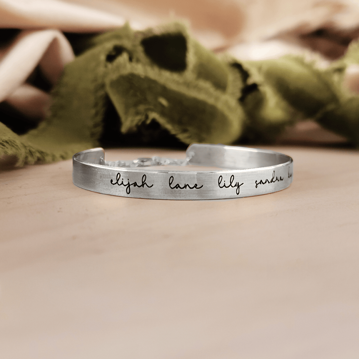 A gold-colored cuff bracelet with custom names engraved on it, displayed on a white surface with a green textured object to the side.
