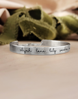 A gold-colored cuff bracelet with custom names engraved on it, displayed on a white surface with a green textured object to the side.
