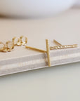 A pair of 14K yellow gold-filled sparkle bar earrings displayed on a wooden surface with a white circular object in the background.