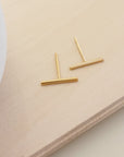 A pair of gold-colored bar earrings placed on a wooden surface next to a white cup.