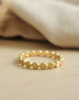 A gold-colored flat beaded stacking ring placed on a wooden surface with a neutral-colored fabric in the background.