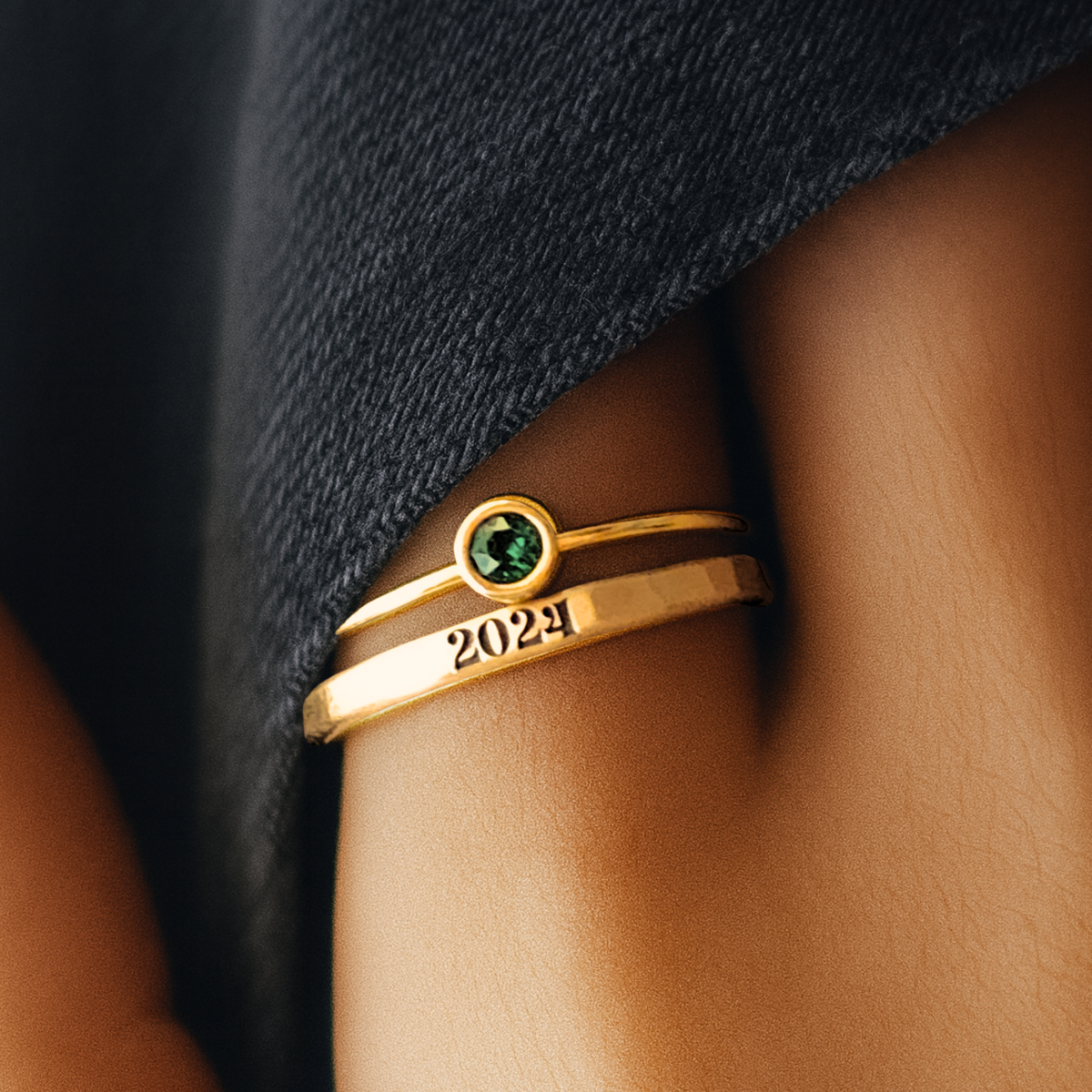 Gold ring with a green gemstone and '2024' on a person's finger against a dark fabric background
