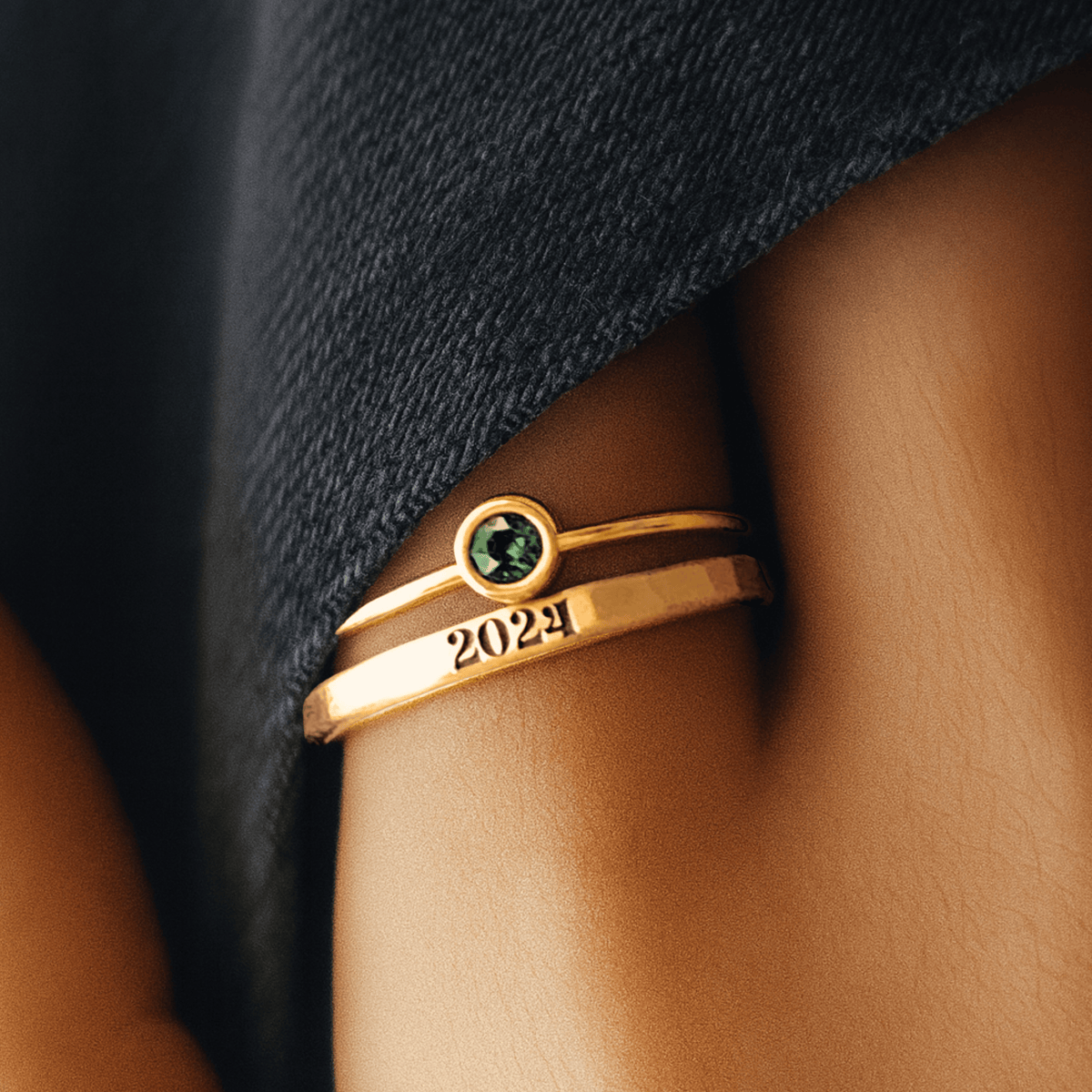 Gold ring with a green gemstone and '2024' on a person's finger against a dark fabric background