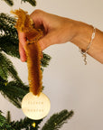 A hand holding a brass ornament with a yellow ribbon and a personalized engraving, attached to a Christmas tree.