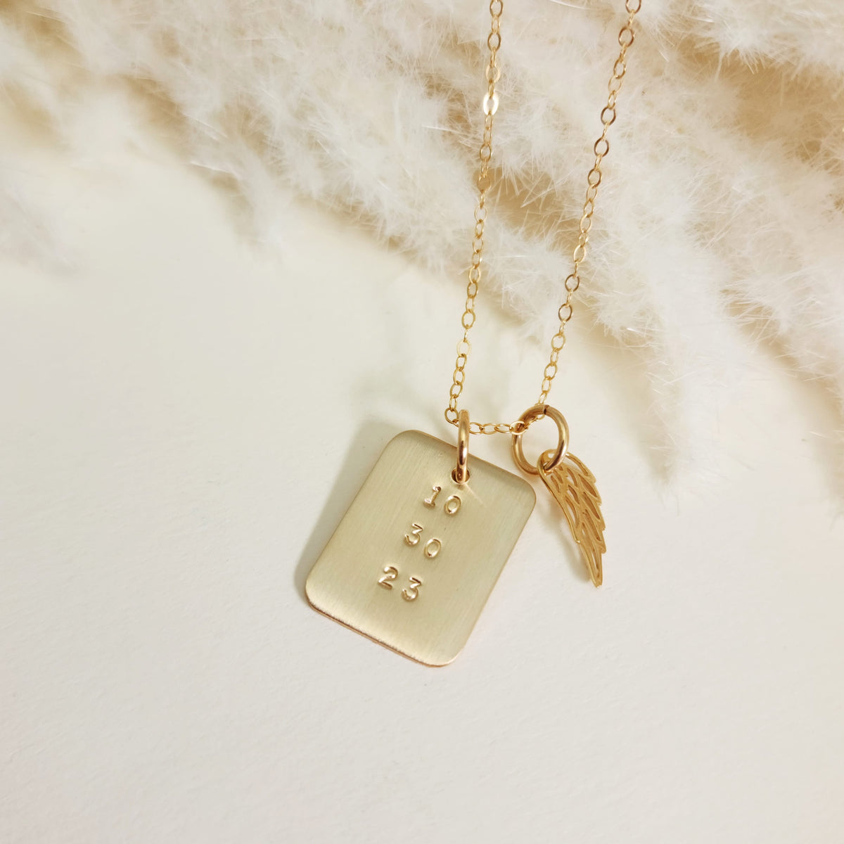 Close-up of gold rectangle date necklace with angel wing charm on soft cream fabric background