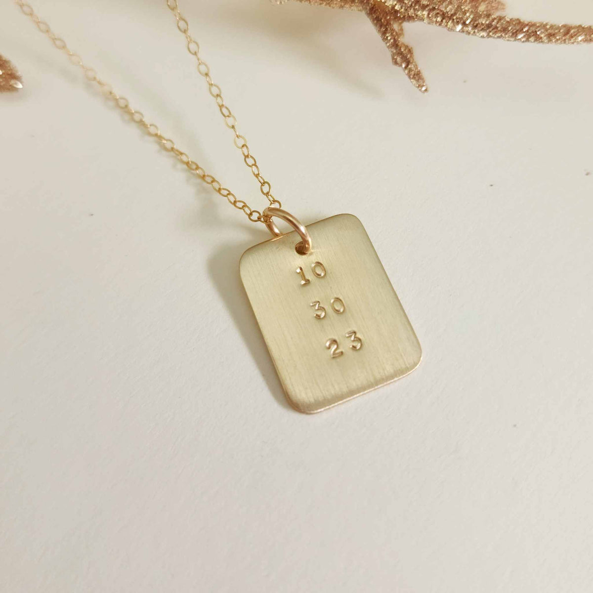 Gold date-stamped pendant necklace on gold chain displayed on a white background with gold leaf accent.