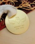 Gold engraved memorial ornament featuring angel wings, halo, and heart design on red and gold holiday fabric.