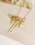 Gold memorial necklace with engraved vertical bar reading “Michael,” gold heart charm, angel wing charm, and birthstone, displayed on wood.