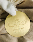 Gold memorial ornament engraved with angel wings, heart, halo, and personalized name and dates on beige fabric.
File Name: gold-memorial-or