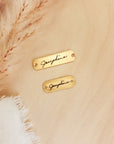 Two gold nameplates with 'Jacqueline' engraved on them, placed on a beige fabric background.