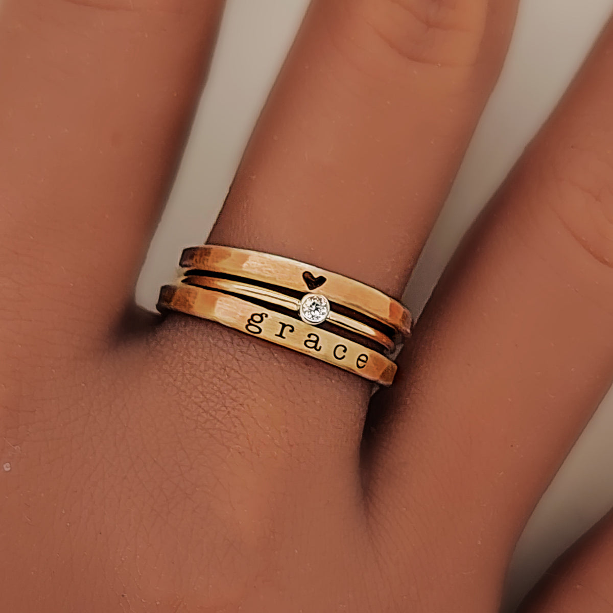 A hand wearing three yellow gold rings stacked on one another, with the top ring featuring a heart, second stone is a clear stone and the word 'grace' engraved on the bottom.
Details