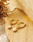 A pair of gold-colored hoop earrings with white freshwater pearl accents placed on a wooden surface with a blurred holiday-themed backdrop.