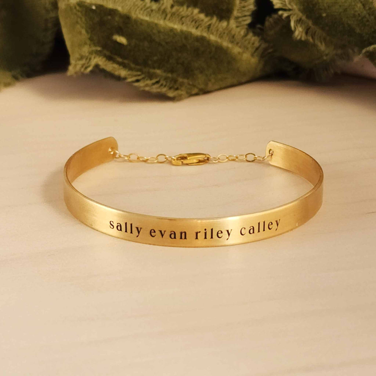 A gold-colored cuff bracelet with custom names engraved on it, displayed on a white surface with a green textured object to the side.