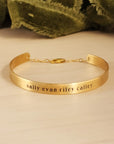 A gold-colored cuff bracelet with custom names engraved on it, displayed on a white surface with a green textured object to the side.