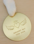 Personalized gold remembrance ornament with engraved angel wings, heart, and halo design on a light wood surface.