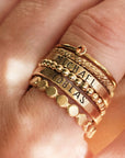 A collection of gold-colored stacking rings with varying designs, some with beaded details and personalized engravings, displayed on a person's finger.