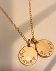 Gold necklace with two round pendants engraved with names on a soft background