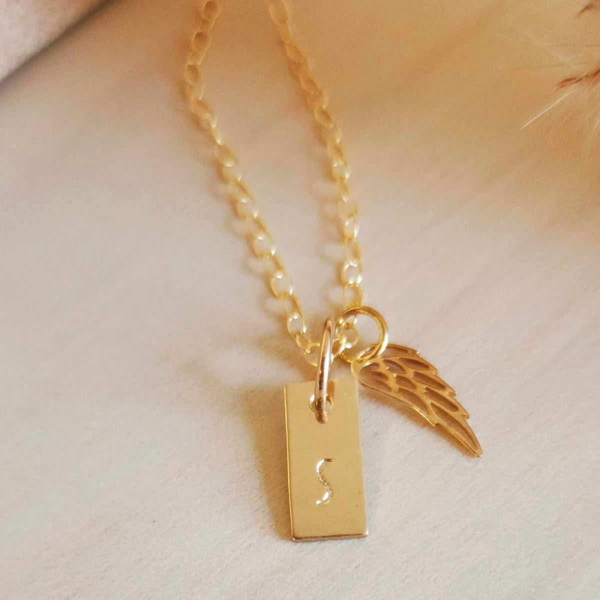 A gold angel wing necklace with a small gold-filled initial tag and a bronze angel wing, displayed against a soft-focus background.