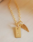 A gold angel wing necklace with a small gold-filled initial tag and a bronze angel wing, displayed against a soft-focus background.