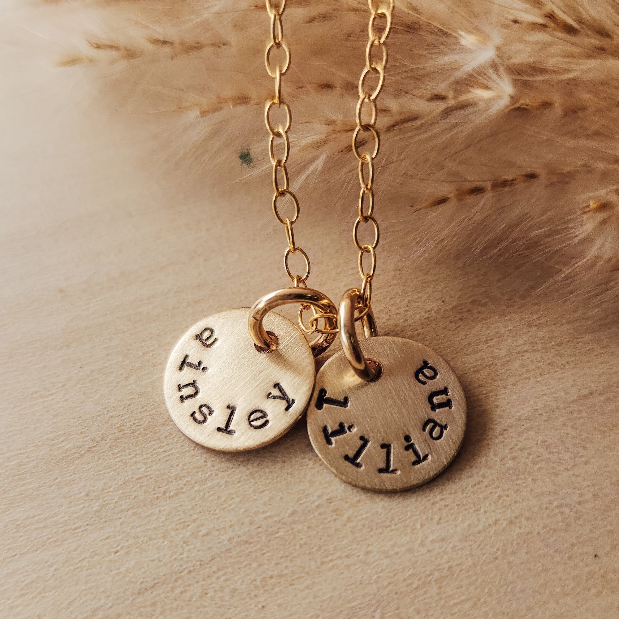 A dainty round name necklace with two gold tags on a chain, laying on a beige surface with wheat in the background.