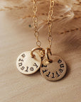 A dainty round name necklace with two gold tags on a chain, laying on a beige surface with wheat in the background.
