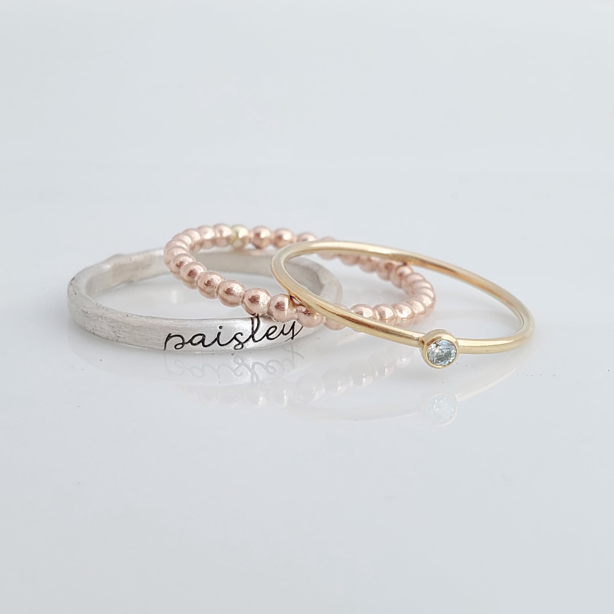 The set includes a silver ring with a brushed texture and engraved in cursive, a delicate rose gold beaded ring, and a thin yellow gold ring with a small bezel-set blue gemstone. The rings are arranged in an overlapping manner, creating a visually appealing composition with soft reflections on the surface below.