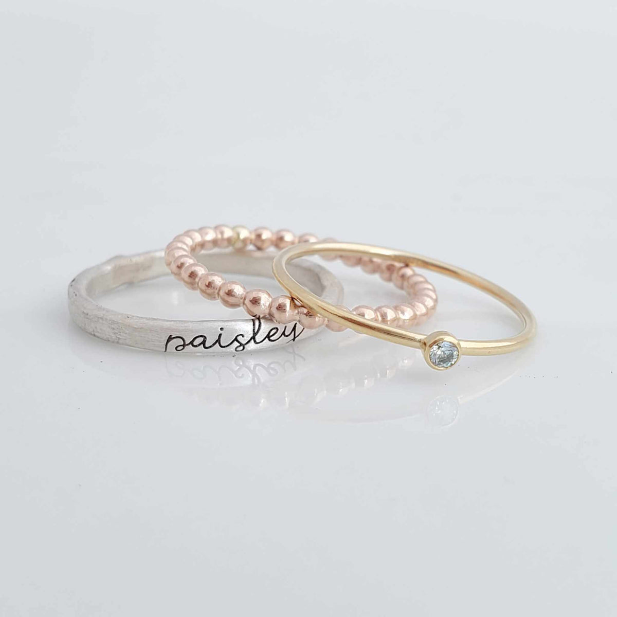 The set includes a silver ring with a brushed texture and engraved in cursive, a delicate rose gold beaded ring, and a thin yellow gold ring with a small bezel-set blue gemstone. The rings are arranged in an overlapping manner, creating a visually appealing composition with soft reflections on the surface below.