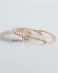 The set includes a silver ring with a brushed texture and engraved in cursive, a delicate rose gold beaded ring, and a thin yellow gold ring with a small bezel-set blue gemstone. The rings are arranged in an overlapping manner, creating a visually appealing composition with soft reflections on the surface below.