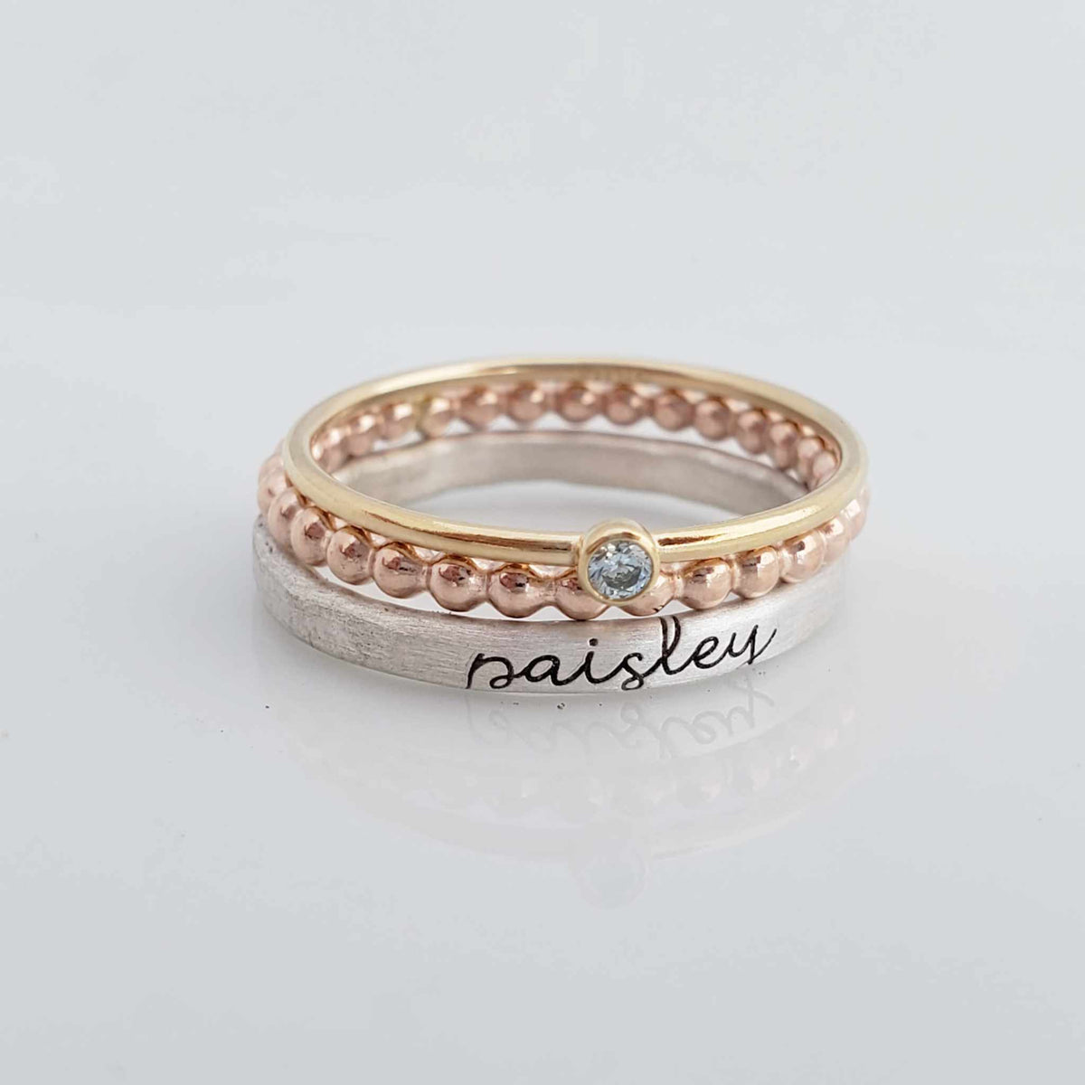 The set includes a silver ring with a brushed texture and engraved in cursive, a delicate rose gold beaded ring, and a thin yellow gold ring with a small bezel-set blue gemstone. The rings are arranged in an overlapping manner, creating a visually appealing composition with soft reflections on the surface below.