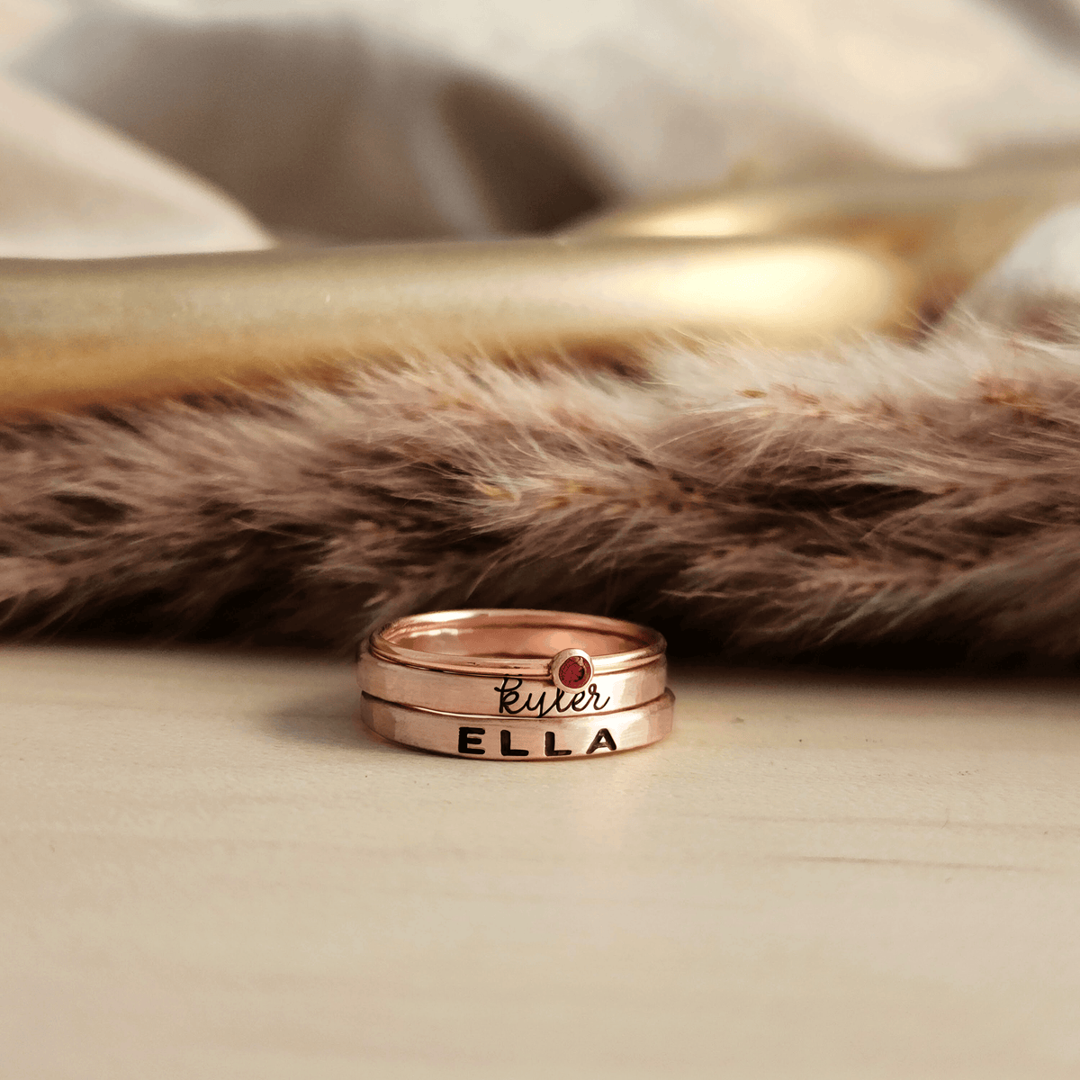 A set of stackable rose gold filled rings, with one clear cz ring and two personalized name rings, displayed on a wooden surface.