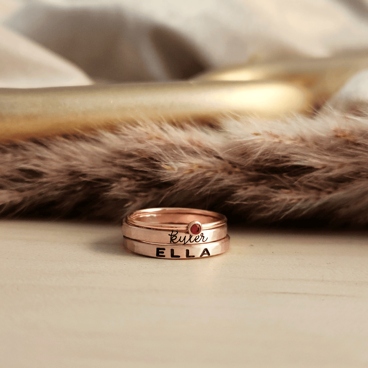 A set of stackable rose gold filled rings, with one clear cz ring and two personalized name rings, displayed on a wooden surface.