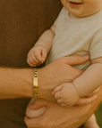 Person holding a baby with a gold bracelet on their wrist, set against a blurred natural background.