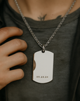 Person wearing a necklace with a silver dog tag on a dark shirt